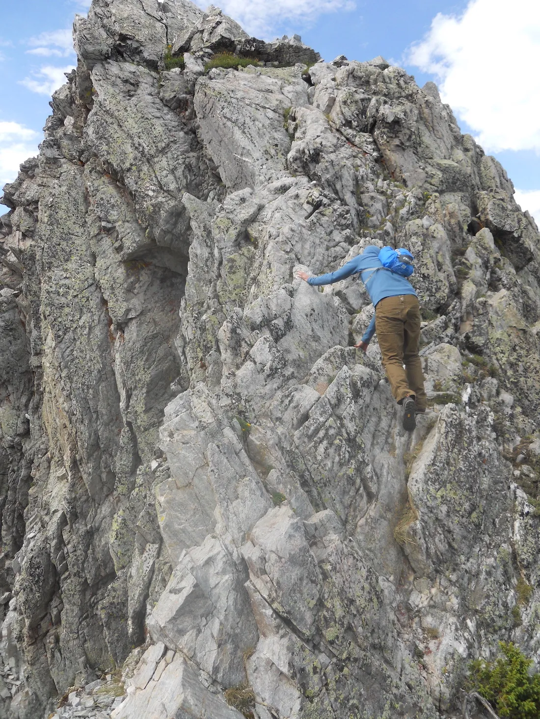 A climber in a blue shirt and pack scrambles up steep, lichen-covered granite on the cirque ridge buttress.