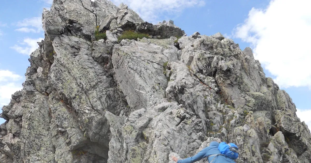 A climber in a blue shirt and pack scrambles up a grey granite buttress on the Quemado Cirque ridge under a partly cloudy sky.
