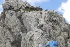 A climber in a blue shirt and pack scrambles up a grey granite buttress on the Quemado Cirque ridge under a partly cloudy sky.