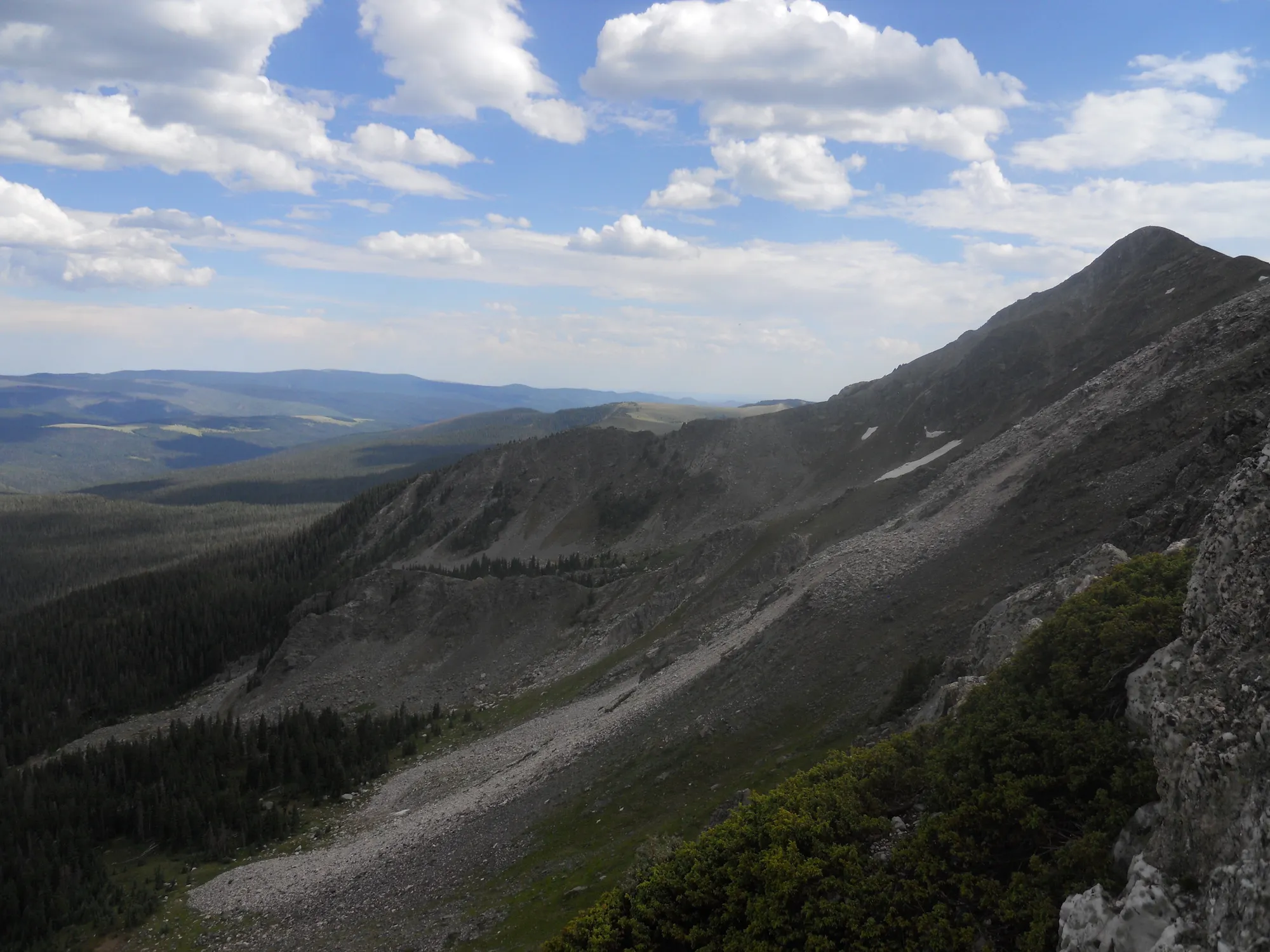 View along the cirque rim toward a grey peak with a long scree slope below it, krummholz in the foreground, and forested valleys under cumulus clouds beyond.